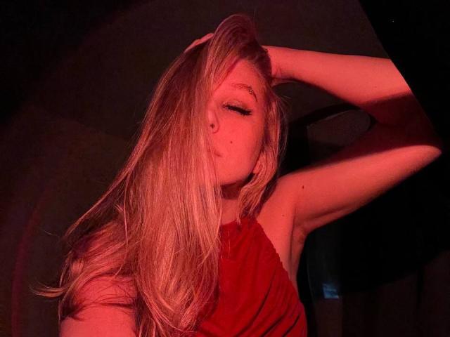Alice, avec de longs cheveux blonds, une robe rouge, le bras levé, les yeux fermés, une lumière rouge, des taches de rousseur sur la joue, un sourire doux, sexy, détendue, attirante..