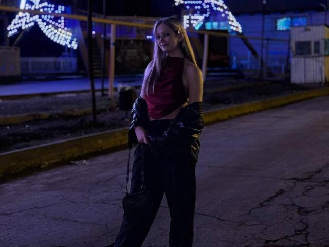 Alice, con el pelo rubio largo, vestida con un top rojo, una chaqueta de cuero negro y pantalones negros, está de pie en una calle agrietada y poco iluminada por la noche, sosteniendo un bolso negro..