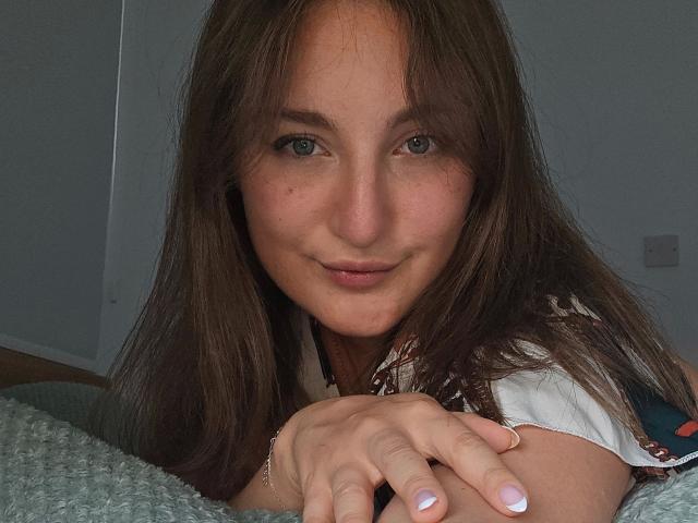 Evelynka-hot with long brown hair, light skin, and blue eyes, smiling slightly, lying on a green textured blanket, hands resting in front. Small nose, natural makeup, pink lips.