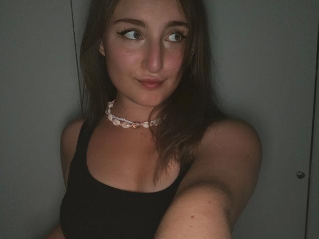 Evelynka-hot with medium tits, wearing a black tank top and pink shell necklace, brown hair, blue eyes, slight smile, taking a selfie in dim lighting.
