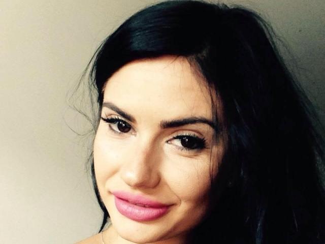 Alexandrina69 has dark, wavy hair, brown eyes, and pink lipstick. She's smiling with long eyelashes, and her skin is fair. Boobs aren't visible; focus is on her face.