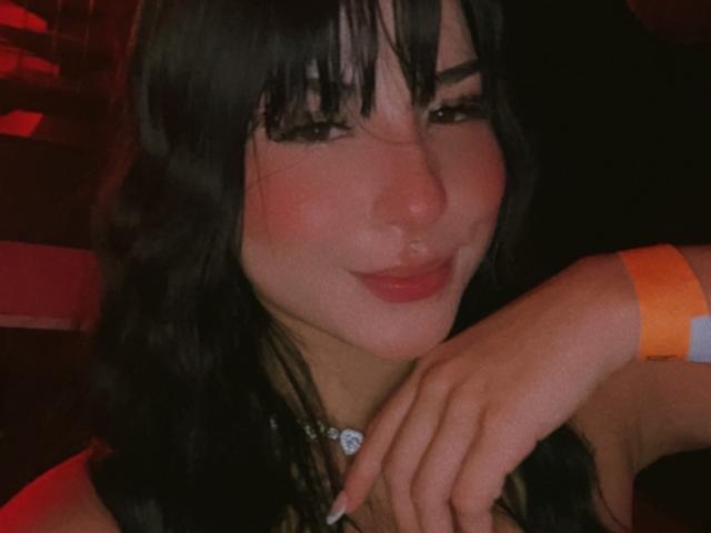Grace Clarke, con i capelli neri, il rossetto rosso e un sorriso adorabile, indossa un braccialetto arancione e ha il mento appoggiato sulla mano, appare sexy e sicura di sé..