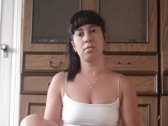 AlinaMiss has black bangs, a white tank top showing her small tits, and a neutral expression, sitting in front of a wooden cabinet with glass doors.