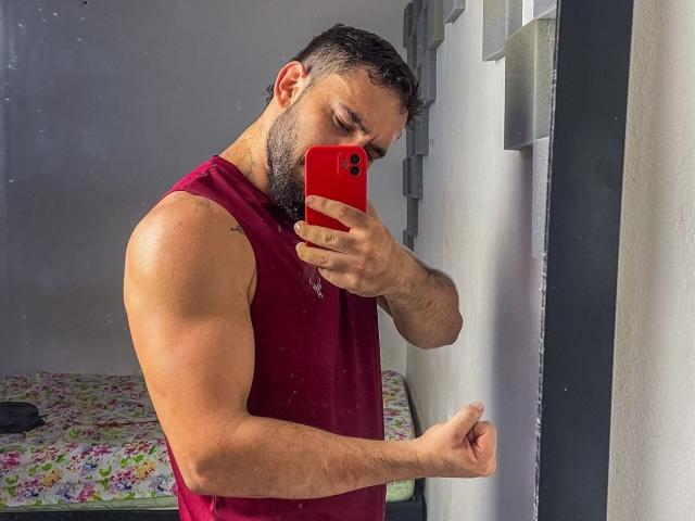 Latinafuckfest, muscular, bearded, wearing red sleeveless shirt, holding red phone, taking mirror selfie, right arm flexed, short dark hair, light skin, standing in front of white wall.
