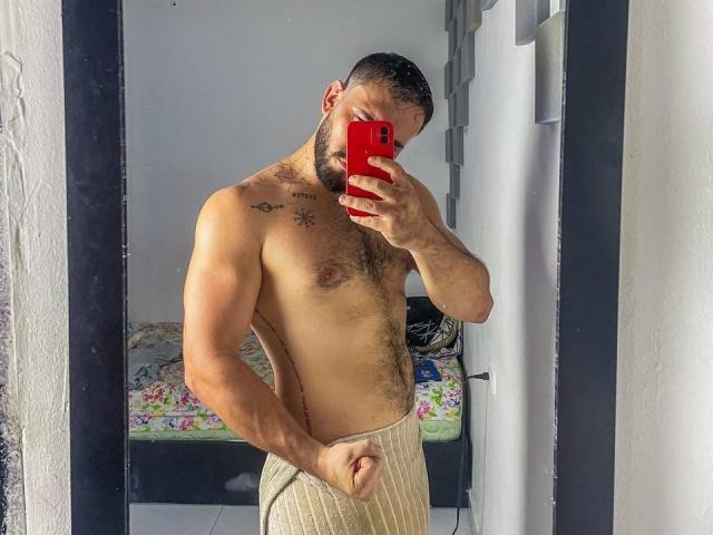 Muscular LatinaFuckFest with short dark hair, beard, chest tattoos, and a towel around his ass, taking a mirror selfie with a red phone in a brightly lit room.