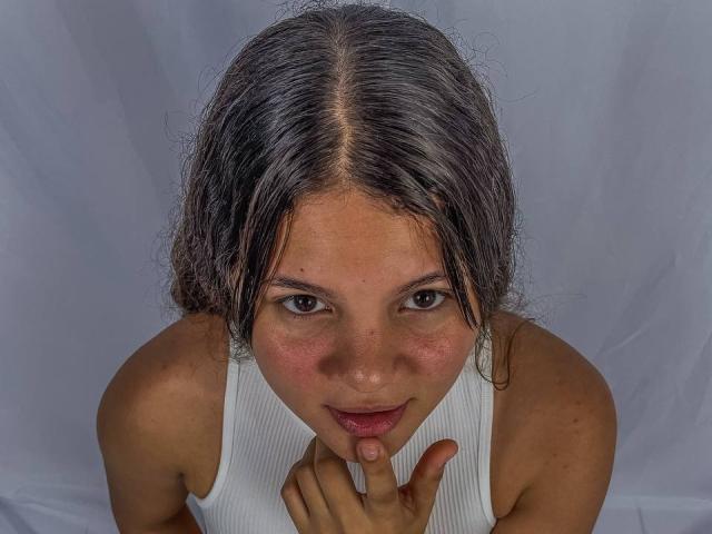 LatinaFuckFest with gray, curly hair, brown eyes, and pink cheeks, wearing a white ribbed tank top, biting her finger with a slightly open, pink-lipped mouth.