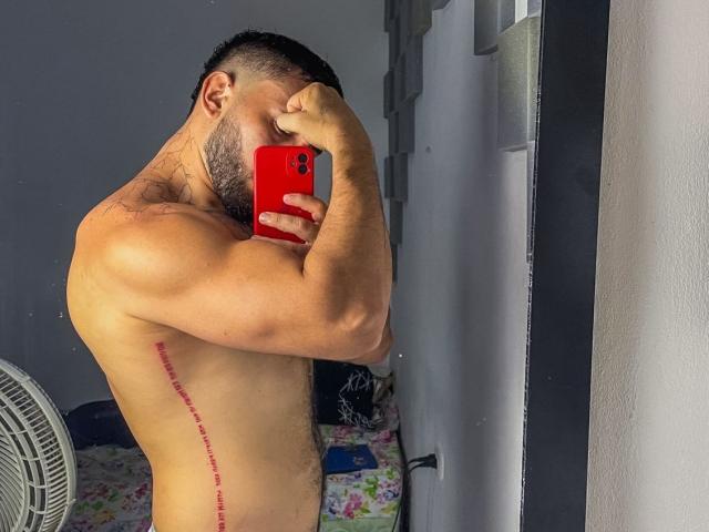 LatinaFuckFest with a muscular, hairy, tan body, red phone, and tattooed side, taking a mirror selfie, covering his face. His cock is not visible.