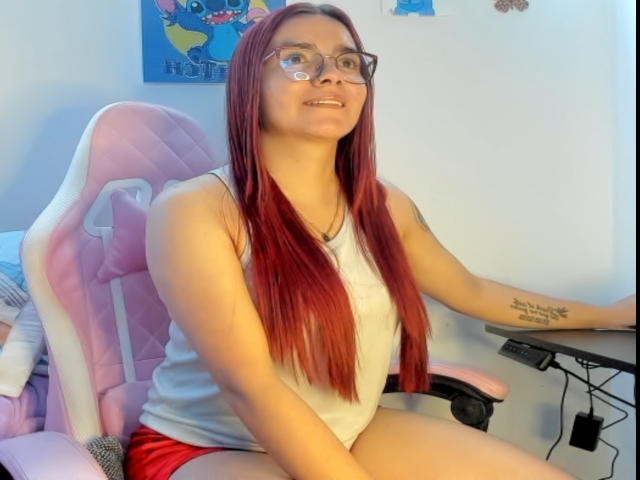 EimmyHoskX with red hair, glasses, white tank top, red shorts, sitting on pink gaming chair, smiling, tattoo on left forearm, light skin, slim, cute, relaxed.