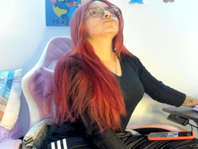 EimmyHoskX with long red hair, glasses, black top, sitting on a pink chair, looking up, right arm on desk, black pants with white stripes, computer in background.