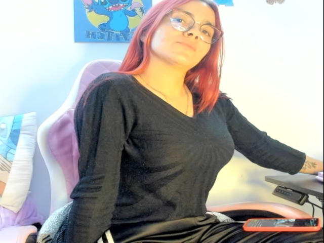 EimmyHoskX with red hair, glasses, black V-neck top, small tits, sitting on a pink chair, black pants, right hand on a keyboard, relaxed expression.