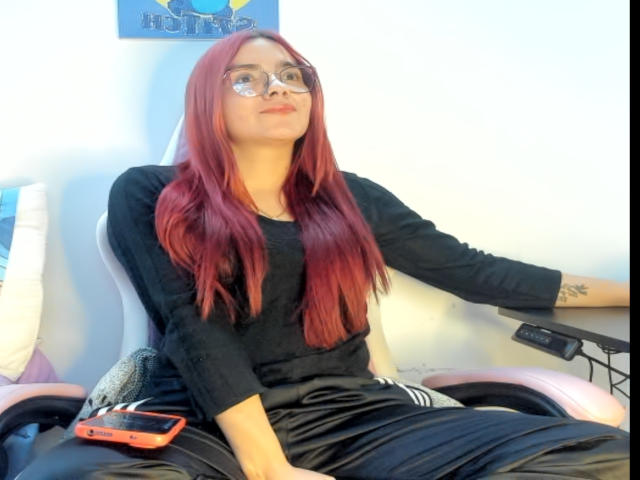 EimmyHoskX with red hair, glasses, black shirt, black pants, sitting on a pink chair, right arm tattoo, red phone on left, relaxed expression, white wall background.