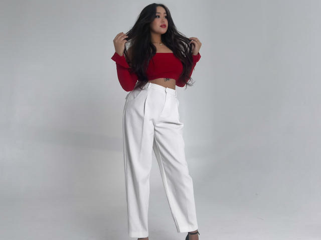 CrystalCharm with long black hair, red off-shoulder top, white high-waisted pants, small tattoo under her right breast, standing, hands in hair, looking at camera, light skin.