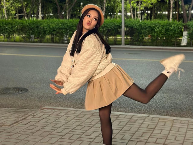 CrystalCharm, with long black hair, pouty lips, beige hat, chunky sweater, tan skirt, black tights, and white boots, kicks one leg up on a sidewalk.