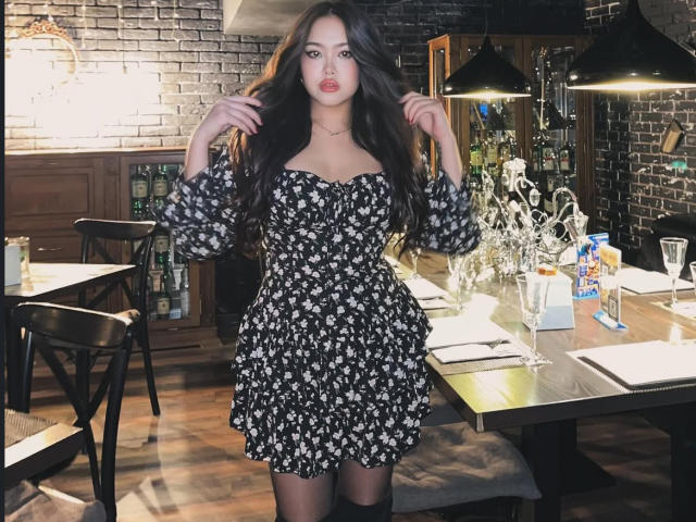 CrystalCharm has long dark hair, wearing a black floral dress, standing in a dimly lit restaurant, hands in hair, black chair to her left, table with glasses and snacks.
