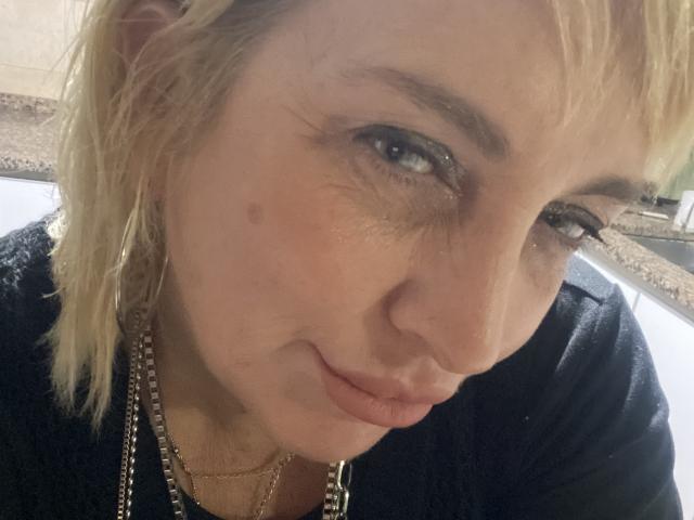 LunaSensualX with blonde, wet-looking hair, blue eyes, slight smirk, black shirt, necklace, small beauty mark on right cheek, close-up selfie, looking down.