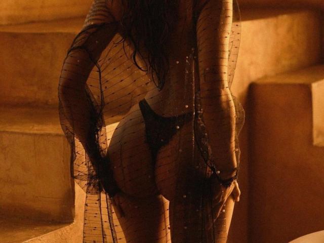ElectroLivviy's back is to the camera, wearing a black mesh robe, exposing her bare ass and black thong, with long dark hair, in warm, golden light.