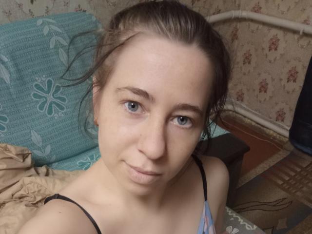AlinaMiss has fair skin, blue eyes, and brown hair in a messy bun. She's wearing a blue and pink top with black straps, showing her small tits.