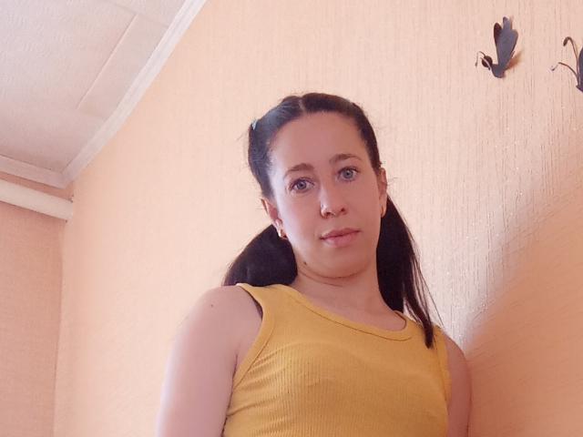AlinaMiss with puffy lips, wearing a yellow ribbed tank top, black pigtails, and blue eyes, standing against a peach wall with black butterfly decor.