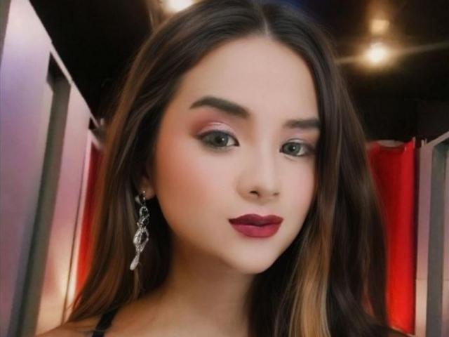 AlluringJhana has long brown hair, dark red lipstick, and wears dangling earrings, with makeup highlighting her eyes and eyebrows. She's looking slightly to the side with a subtle smile.