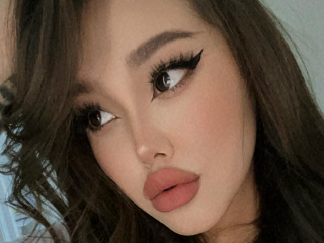 CrystalCharm's face with pouty pink lips, thick black eyeliner, and wavy brown hair, looking to the side with soft, fair skin and slightly flushed cheeks.