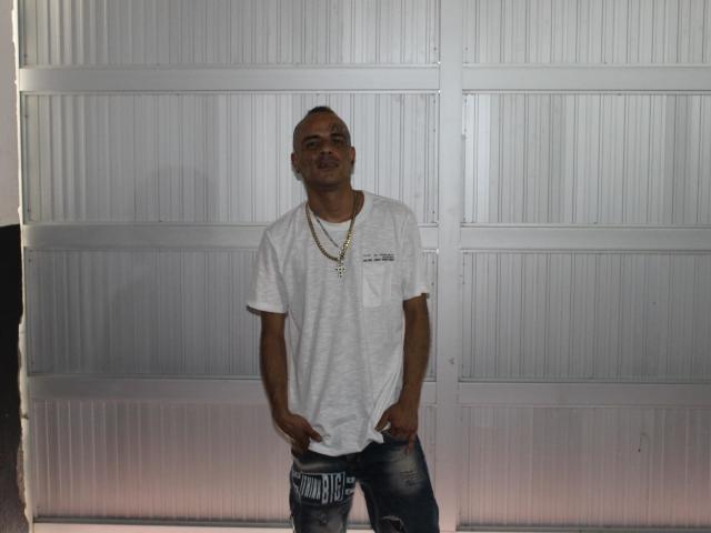 StrongAlan, bald, wearing a white tee and ripped jeans, hands in pockets, gold chain, smiling, standing in front of a white, ribbed, metal door.