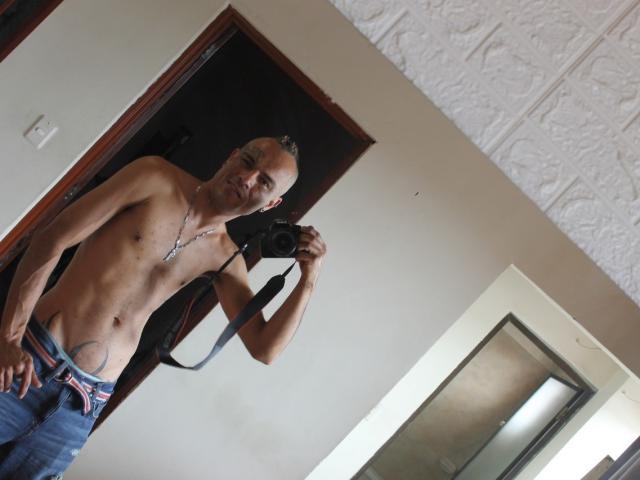 Shirtless StrongAlan with a camera, holding it in his right hand, wearing blue jeans with a visible "Pride" waistband, standing in a room, smiling.