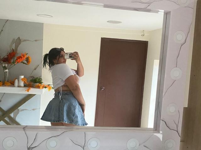 EmmaMyles with a curvy body, white t-shirt, and blue denim skirt, taking a mirror selfie, showing her ass, brown hair in a ponytail, holding a phone.