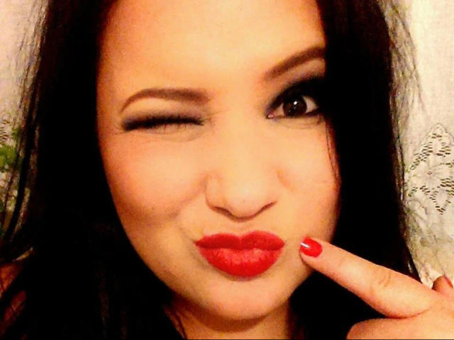 LaVa24-hot a les cheveux noirs, cligne un œil et fronce les lèvres rouges avec un doigt. Elle a du vernis à ongles rouge, et une expression mignonne et espiègle..
