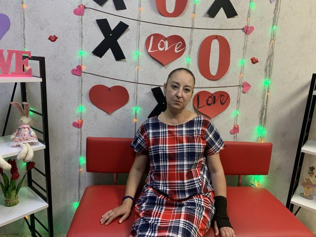 MelissaShell, bald, wearing a red, white, and blue plaid dress, black wristband, sitting on a red bench, surrounded by heart and "X" decorations with green and orange lights.