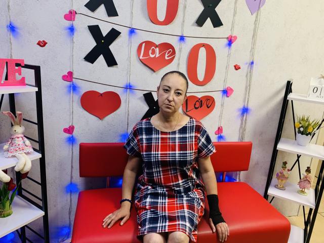 MelissaShell, bald, wearing red and blue plaid dress, black wristband, sitting on red bench, surrounded by heart and love decorations, blue fairy lights, white shelves with dolls.