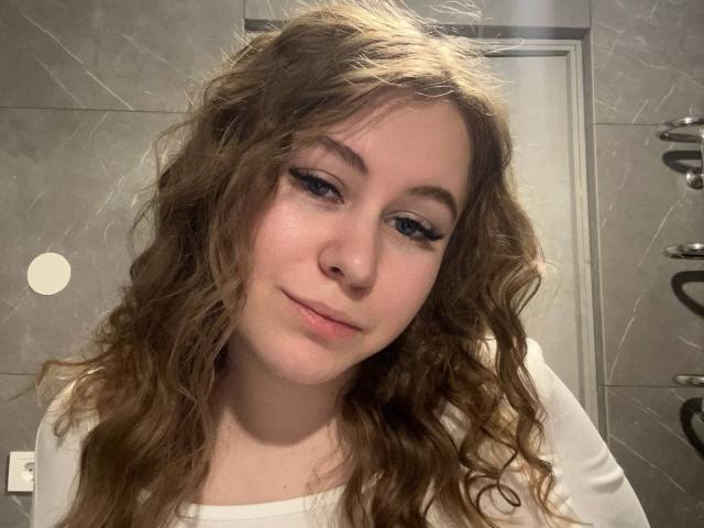 Ariashka with wavy brown hair, light skin, and winged eyeliner, wearing a white shirt, smiling softly at the camera. Her ass is not visible.