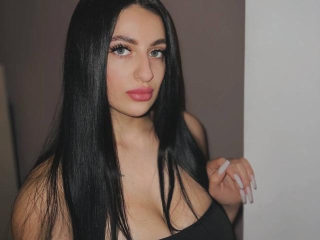 ArielGemme with long black hair, green eyes, and pink lips, wearing a black top, showing her tits, holding a white object, standing against a white wall.