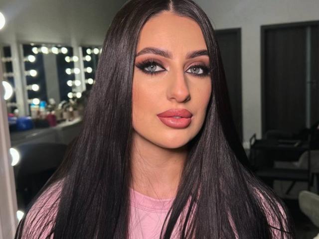 ArielGemme has long black hair, full pink lips, and dramatic eyeliner, wearing a pink top, in a brightly lit mirror room, with a serious, slightly pouty expression.