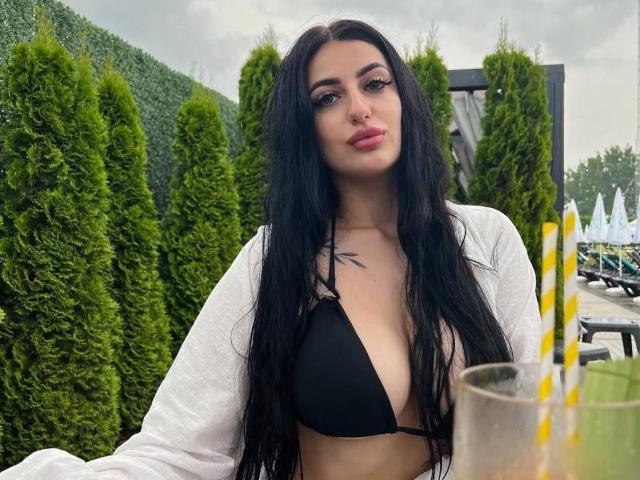 ArielGemme with long black hair, wearing a black bikini top showing her small tits, white cardigan, and pink lipstick, sitting outdoors with a drink and striped straw.