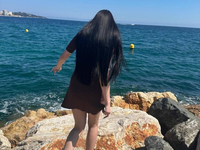 ArielGemme with long black hair stands on rocky shore, wearing a brown dress, arms out, facing blue ocean, yellow buoys, and clear sky. Her ass is visible.