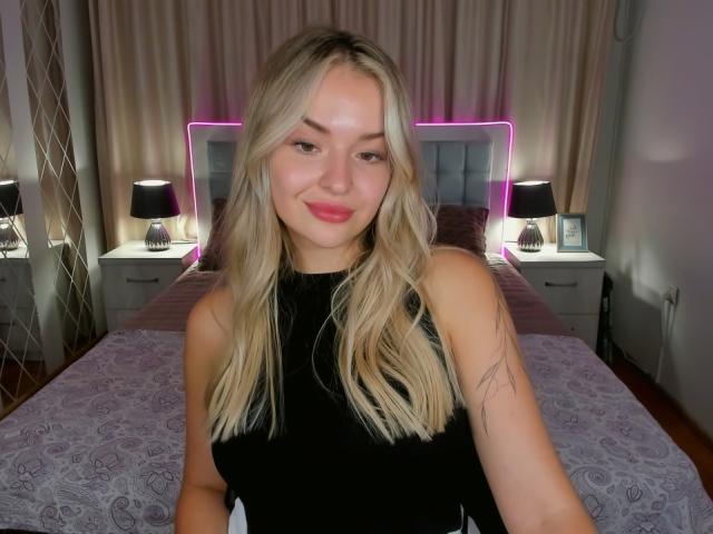 EvaAdderiy, loira, com lábios rosados, vestindo uma blusa preta sem mangas, com uma pequena tatuagem no braço direito, está em pé em frente a uma cama com cabeceira roxa neon, luminárias pretas e um cobre-leito cinza..
