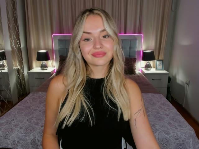 Eva Adderiy, con cabello rubio, top negro sin mangas, labios rosados, un tatuaje pequeño en el brazo derecho, sonriendo a la cámara, cama con luces de neón moradas en el fondo..