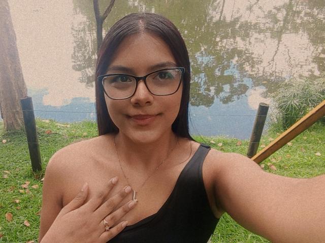 EmilyRobeerts with black glasses, medium brown skin, long dark hair, small tits, black sleeveless top, hand on chest, subtle smile, standing by a reflective water surface.