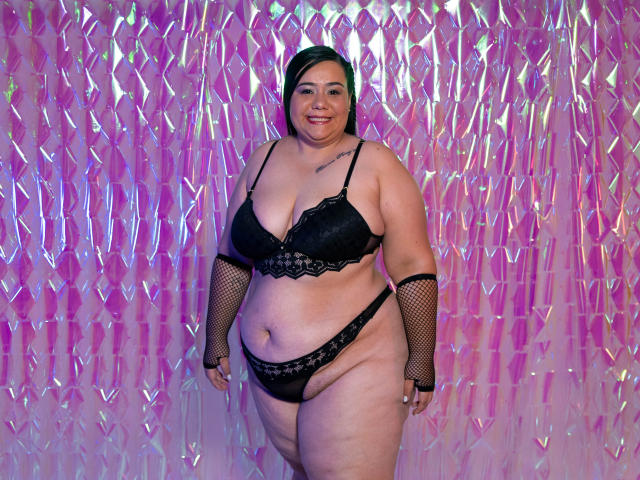AndreaaX69, plus-sized woman with black lace bra and panties, fishnet arms, green hair, smiling, tattoo on chest, standing, pink and purple neon background.