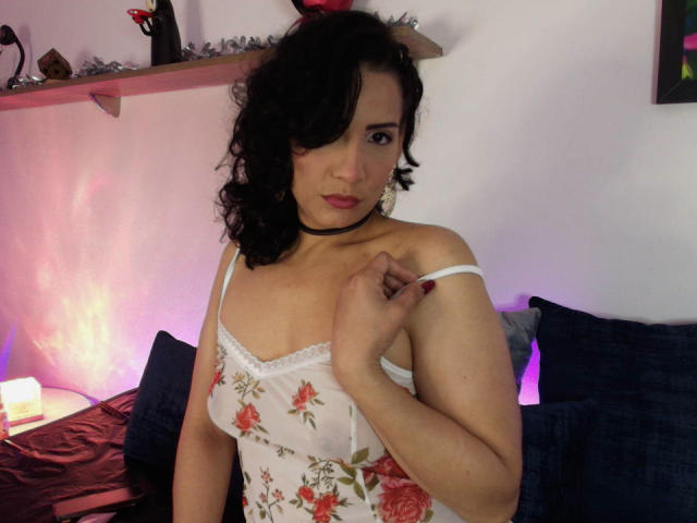 EllieNova with curly black hair, wearing a floral white camisole, pulling down the strap to show her small tits, has pink lipstick and a black choker, sitting on a bed.