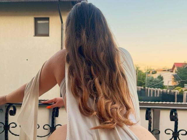 AzzyIce mit langen, welligen braunen Haaren, vom Rücken zur Kamera, trägt ein weißes, ärmelloses Oberteil und sitzt auf einem Balkon, die Hände auf der Reling, der Hintern ist sichtbar, orangefarbene Nägel..