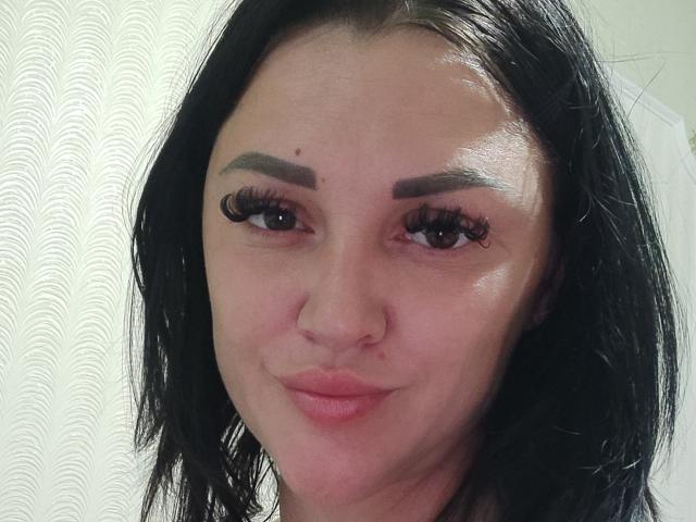 XCrazyBabeX has dark brown eyebrows, long black eyelashes, and a small beauty mark on her left cheek. She's smiling with slightly pinked lips and black shoulder-length hair.
