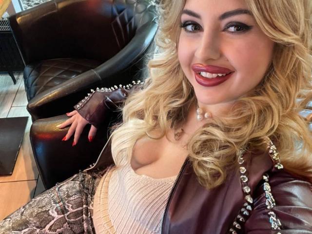 GalaxyNatalie met blonde krullen, rode lippenstift, een parelketting, decolleté in een roze top, een bruine leren jas, broek met slangprint, rode nagels, zittend in een zwarte leren stoel..