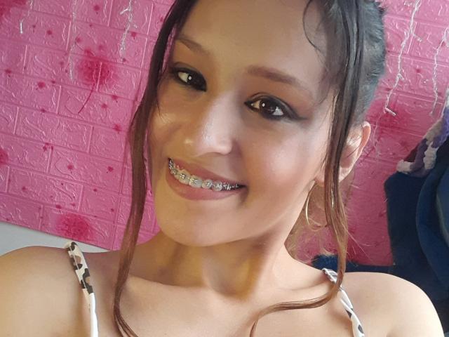 WorthyOfBdsm has brown eyes, a smile showing metallic braces, and wears a leopard print top. She has hoop earrings and her dark hair is in a messy updo.