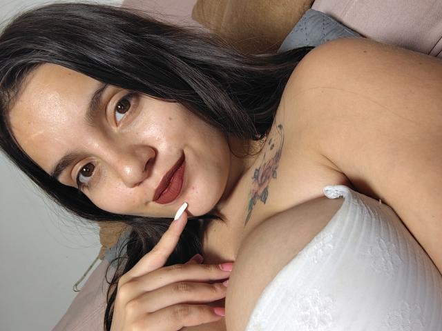 ShiaraSexy with dark hair, brown eyes, and red lipstick, showing off her large tits in a white lace bra, touching her lip with a finger, tattoo on her chest.