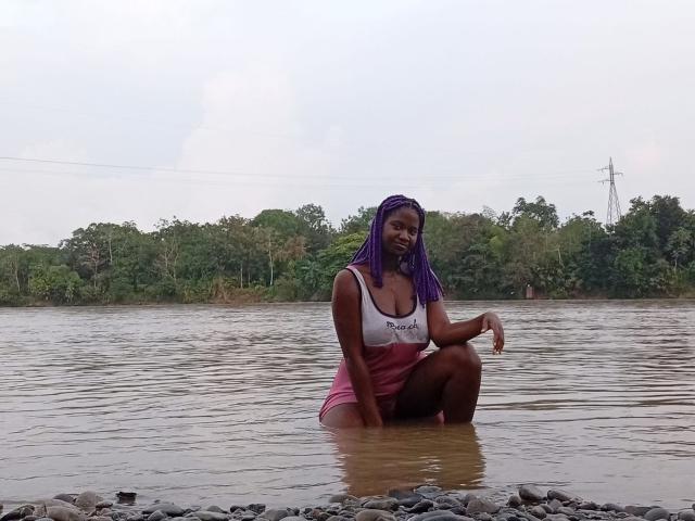 VioletaWal with dark skin and purple braids, kneeling in a river, wearing a white tank top and pink shorts, showing her tits and ass, with a calm water background.