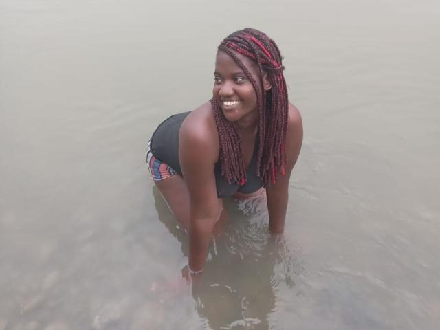 VioletaWal is kneeling in water, smiling with dark skin and red braided hair, wearing a black top and plaid shorts, showing her ass and small tits.