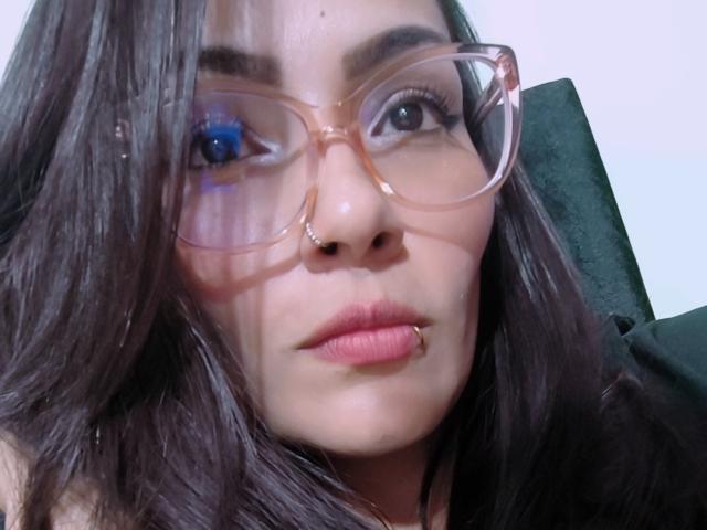AliceGiorgia with brown hair, blue eyes, nose ring, and glasses, has pink lips and a lip piercing, looking serious. Her tits aren't visible. She's wearing a black top.