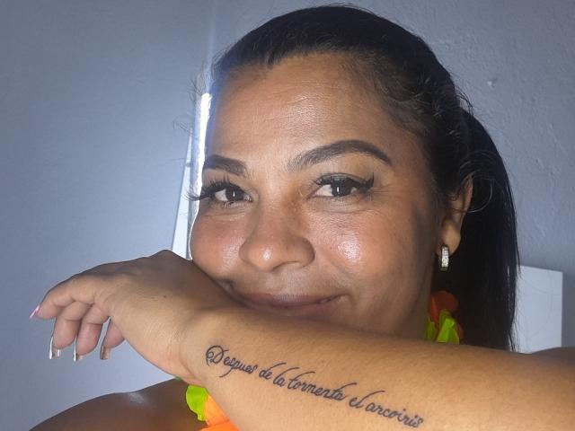 BrunetteScarlet's face with dark hair, brown eyes, and a tattoo on her right forearm reading "Siempre de la mano de Dios" in cursive. She's smiling, wearing a yellow-orange necklace.
