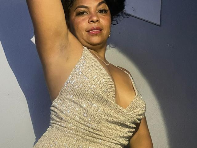 BrunetteScarlet with dark curly hair, wearing a sparkly, low-cut, beige dress, exposing her tits, arm raised, smiling, light skin, standing in shadowed light.